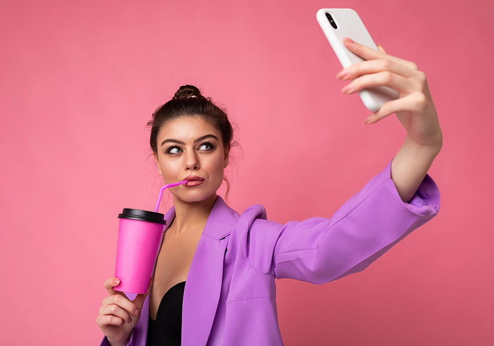 An influencer is taking a selfie with a pink cup and phone.