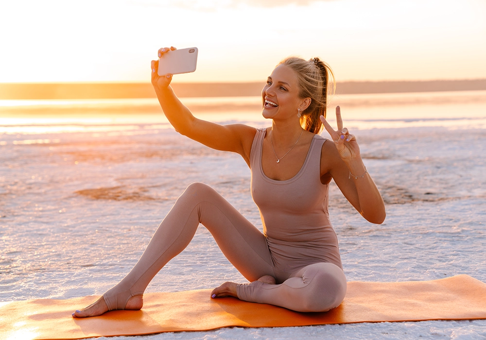 An influencer is taking a selfie while sitting on a yoga mat on the beach.