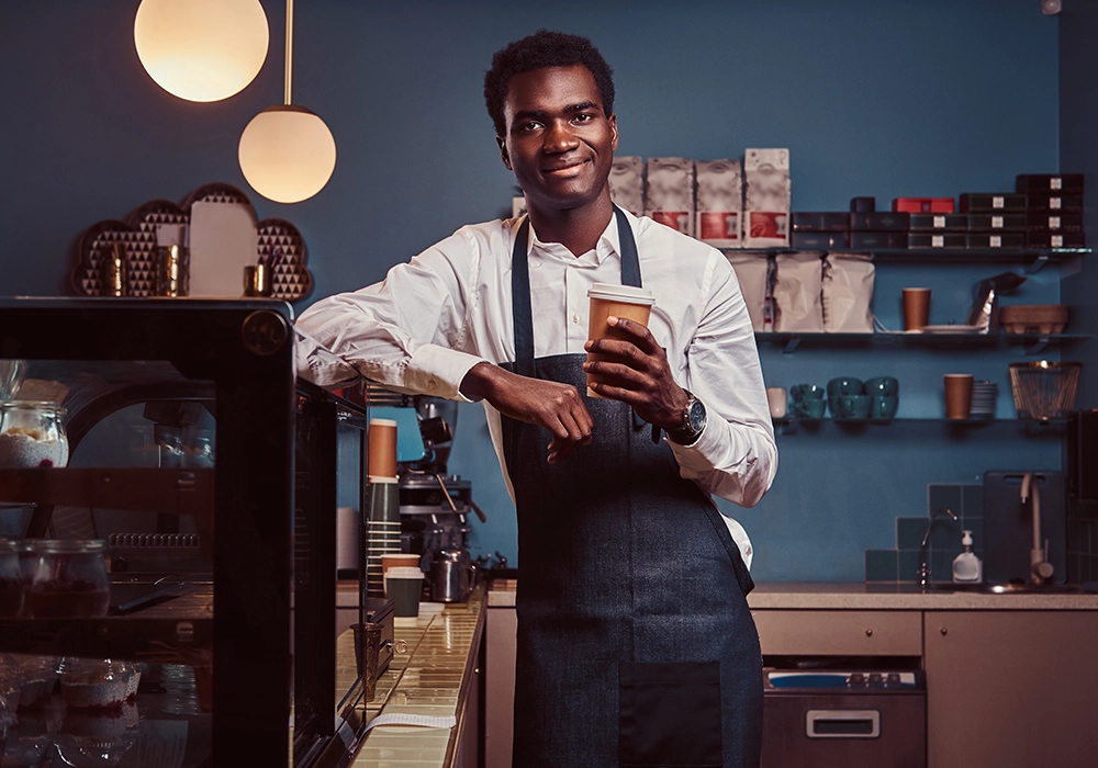 Barista, endorsed by a celebrity, smiling at the camera while holding a takeaway Starbucks coffee cup in a café setting.