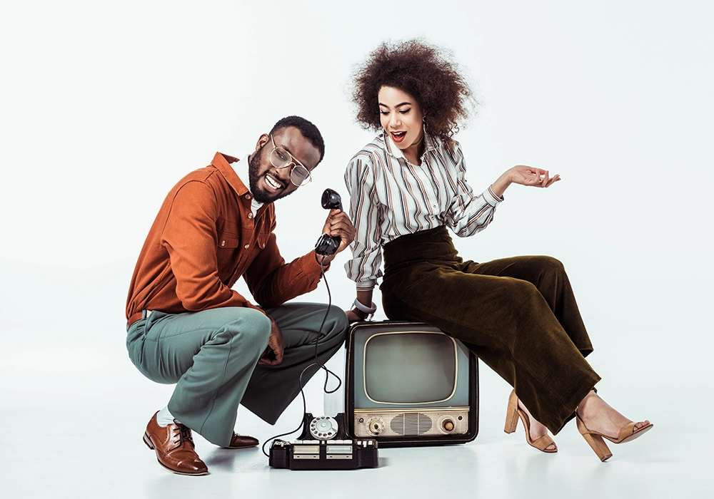 A black man and woman sitting on a white background with an old tv in a traditional advertising setting.