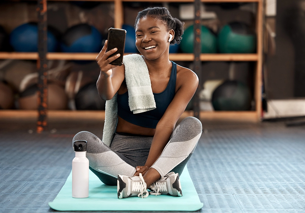 A smiling woman, embodying the Social Proof Theory, sits on a yoga mat taking a selfie at the gym, with a towel around her neck and a water bottle beside her.