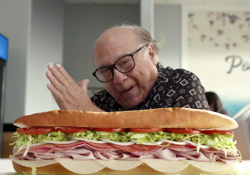 Danny DeVito smiling behind a large Subway sandwich, appearing pleased with the celebrity endorsement.
