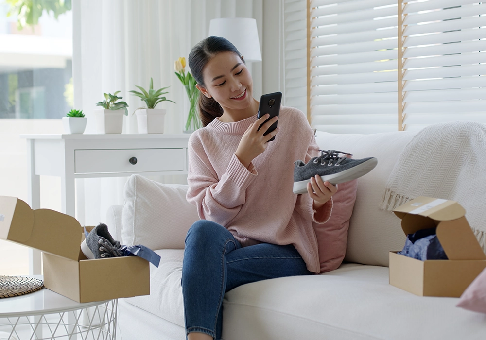 A woman sitting on a couch looking at her phone while admiring the benefits of various shoes.