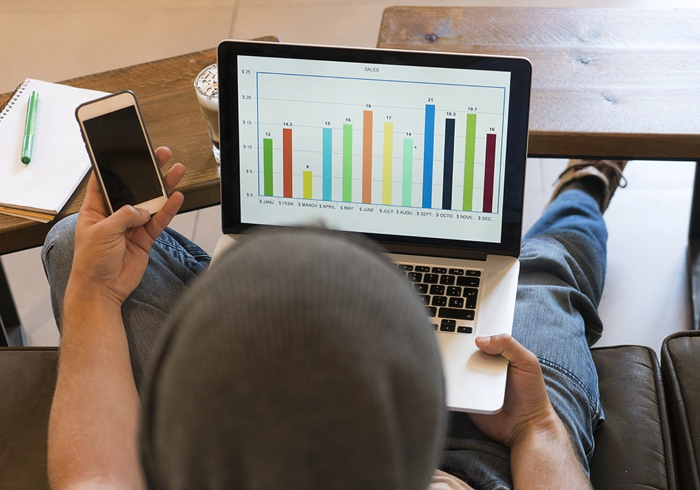 Person holding a smartphone and looking at a typical conversion rate sales graph on a laptop screen while sitting at a wooden table.