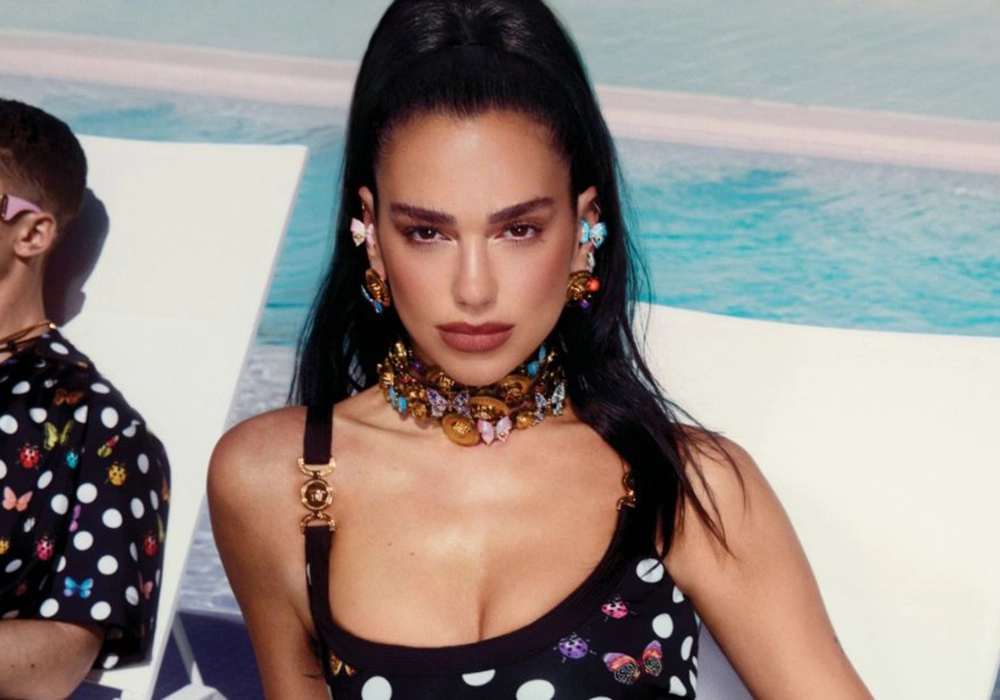 Dua Lipa with sleek hair wearing a polka dot dress and power of celebrity-inspired chunky jewelry, sitting by a pool.