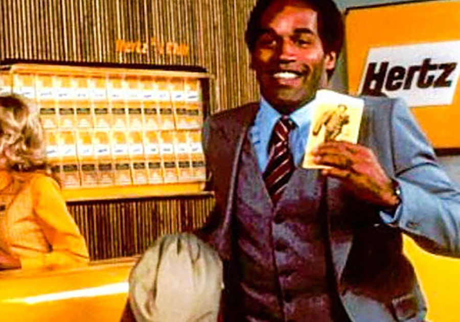 Man in a suit holding a promotional card, smiling, with a yellow car and hertz logo in the background. This image is part of a series on Celebrity Endorsements Gone Wrong.