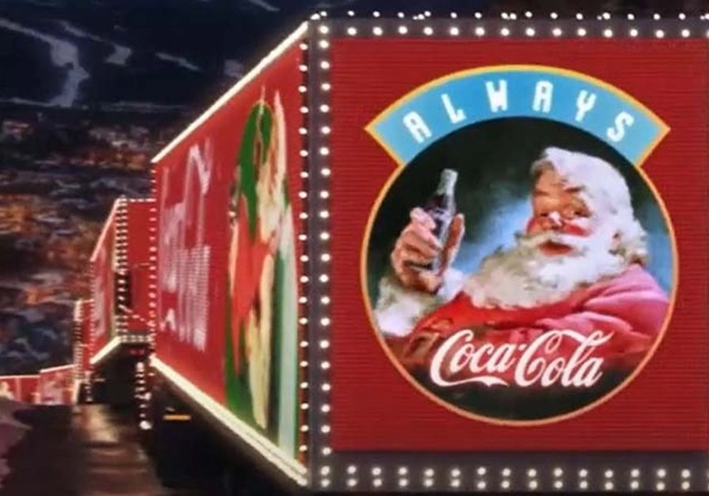 Illuminated Coca-Cola holiday truck, part of the Coca-Cola Campaigns, with an image of Santa Claus holding a Coke, driving by a night-time cityscape backdrop.