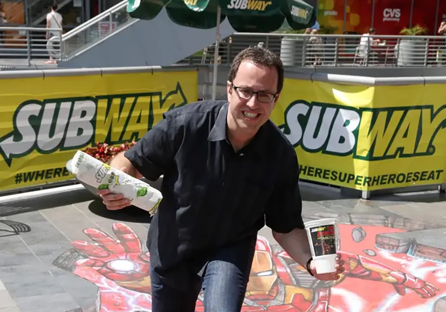 Jared-Fogle in glasses and a black shirt, possibly involved in celebrity endorsements gone wrong, holding a subway sandwich and drink, appears to be tripping over a 3D street art of a superhero scene
