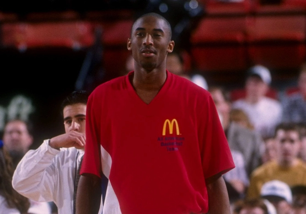 A young basketball player wears a red McDonald's All-American Game shirt, featuring celebrity endorsements gone wrong, standing on a court with spectators in the background.