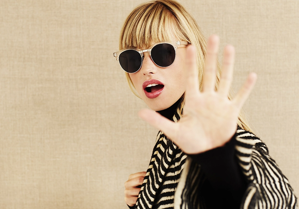 Actress with bangs wearing sunglasses and a striped jacket, extending her hand forward in a stop gesture, epitomizing celebrity endorsements gone wrong.