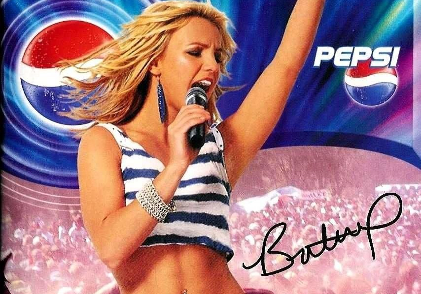 Britney Spears performer sings into a microphone energetically on stage at a Pepsi-sponsored event, surrounded by a large cheering crowd.