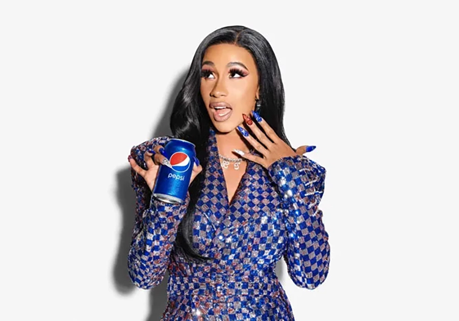 Cardi B, wearing a blue and red patterned dress, holds a can of Pepsi for a celebrity endorsement and expresses surprise with her hand raised to her face.