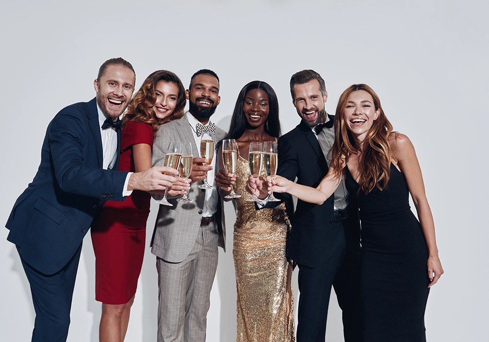 Group of six celebrities in formal wear toasting with champagne glasses against a white background, smiling joyfully at the camera.