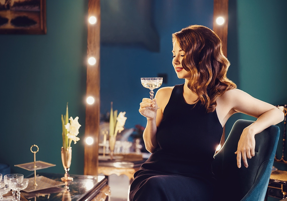 actress in a black dress sitting in a lounge, smiling and holding a cocktail glass, with illuminated vanity mirrors in the background for an TV advertisement. 
