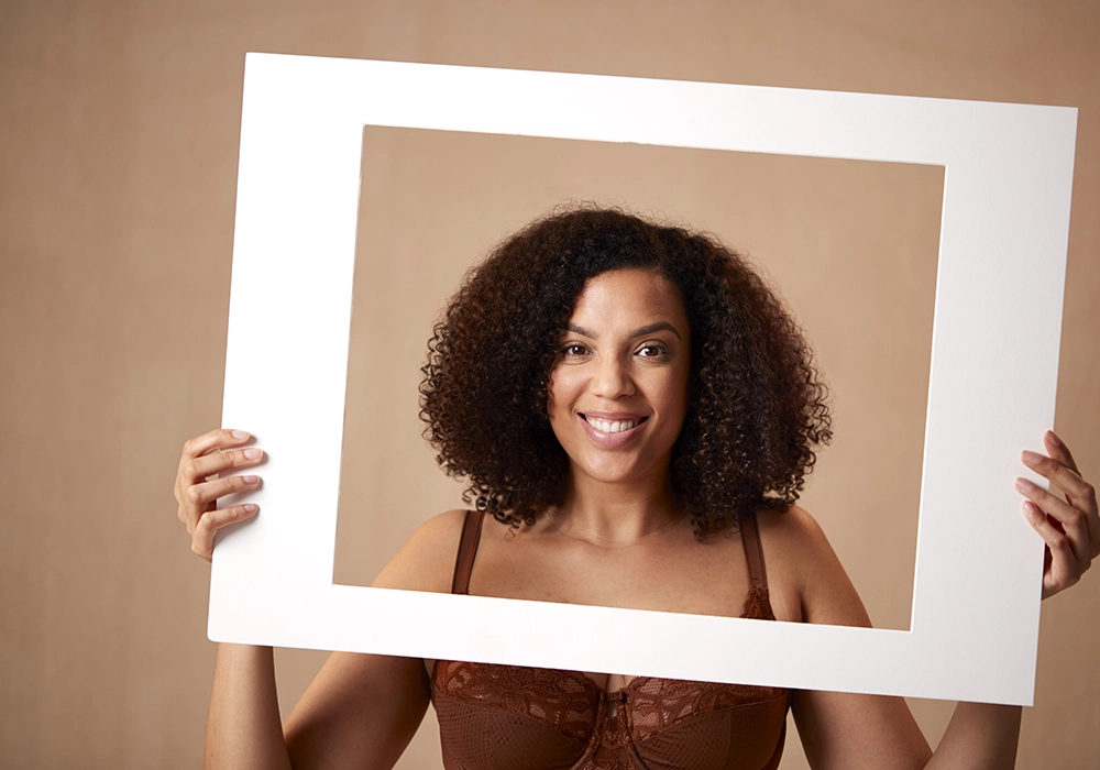 A woman with curly hair smiling and holding a white frame around her face, exuding the power of celebrity against a beige background.