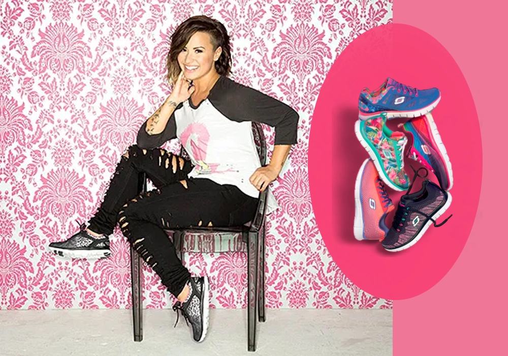 Demi Lovato is sitting on a chair against a floral background, smiling and touching her chin, with an inset image of colorful sneakers from celebrity partnerships on a pink circle to the right.