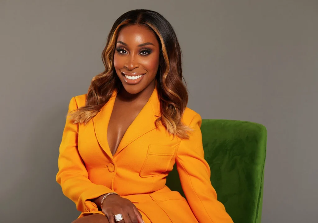 Jackie Aina smiling, dressed in a bright orange blazer, seated on a green chair against a gray background, epitomizing the power of influencers.