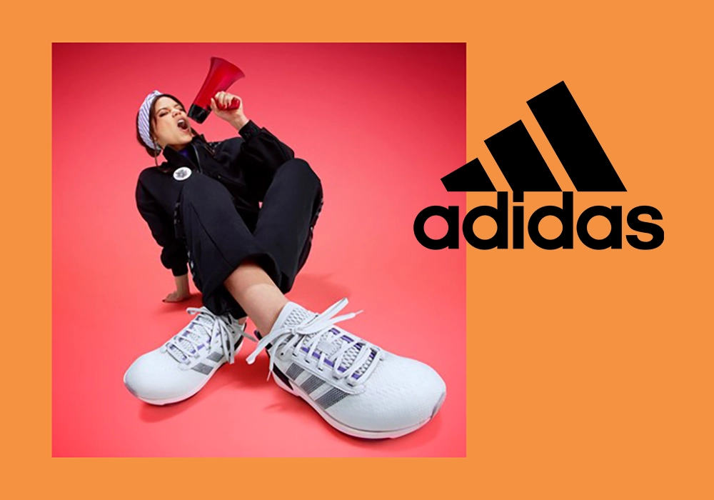 Jenna Ortega in sporty attire holding a megaphone, sitting next to Adidas sneakers, against a coral background.