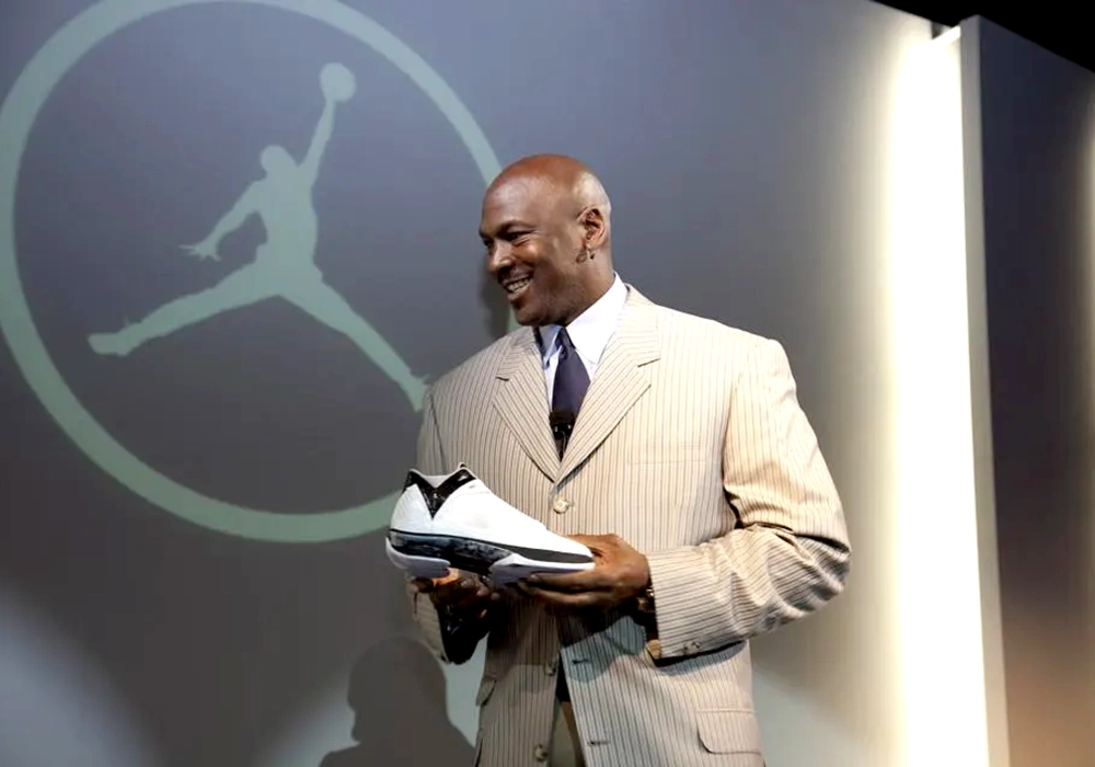 Michael Jordan in a pinstripe suit smiling as he examines a basketball shoe, standing in front of the large Nike logo. He is arguably the most successful Nike celebrity endorsement player of all times.