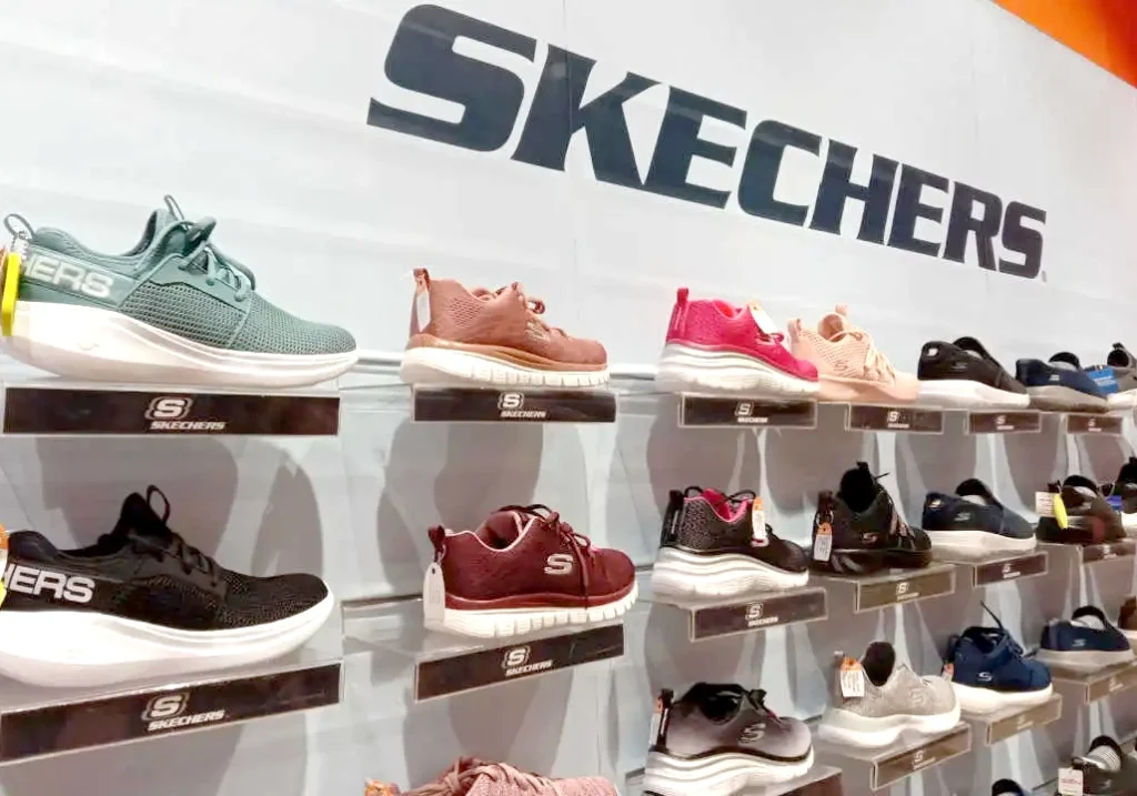 A display of various Skechers shoes, including celebrity partnerships collections, on shelves in front of a large "Skechers" sign on the wall.