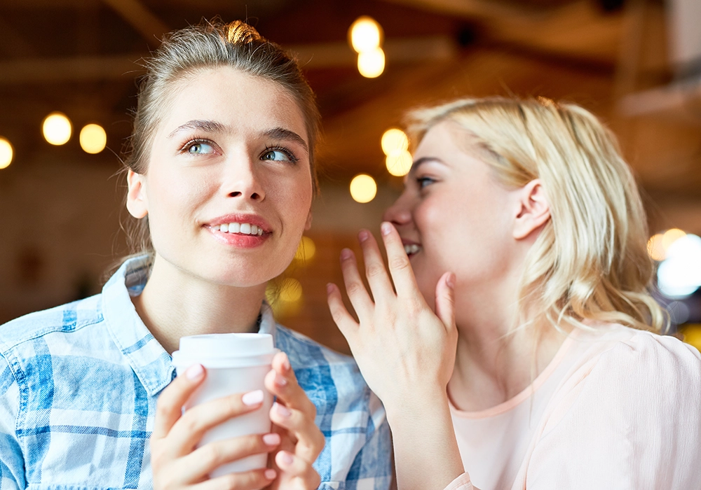 Two women at a cafe, one whispering to the other who is holding a coffee cup and looking away thoughtfully in what seems like an engaging social proof ad.