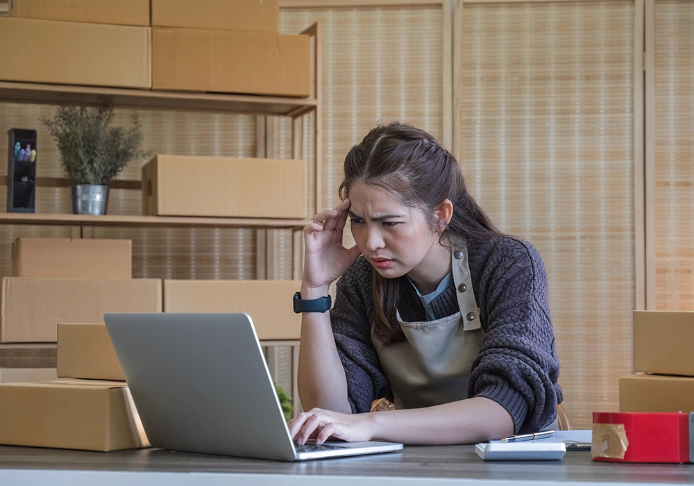 A woman in an apron looks frustrated while using a laptop after experiencing bad customer service in a room filled with cardboard boxes and packing materials.