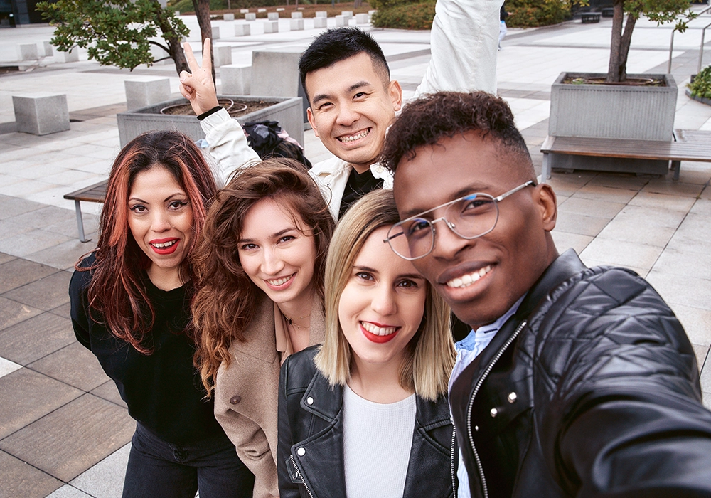 Five diverse friends taking a selfie together outdoors, smiling at the camera, with one person playfully gesturing a peace sign as a form of social proof.