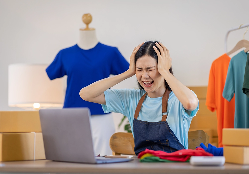 A frustrated woman in an apron covers her ears with her hands in front of a laptop, surrounded by clothing and boxes in a room, distressed by bad customer service.