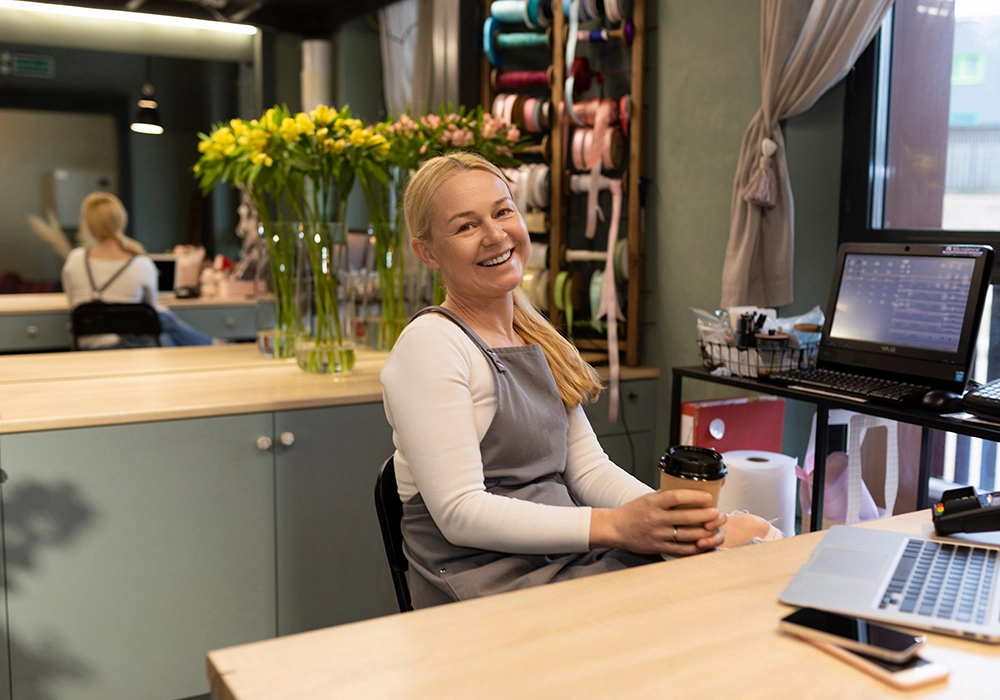A woman in a grey apron smiles at the camera, sitting at a desk with a laptop and a coffee cup in a colorful workspace despite experiencing bad customer service.