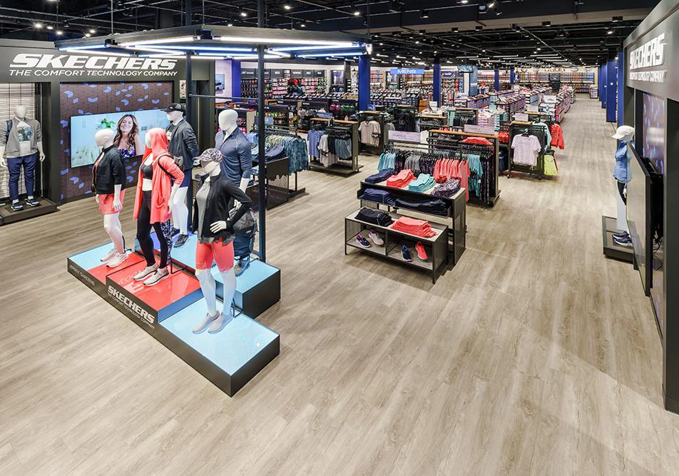 Interior of a Skechers retail store showing various footwear displayed on shelves, mannequins clad in sportswear from celebrity partnerships, and vibrant branding signs.