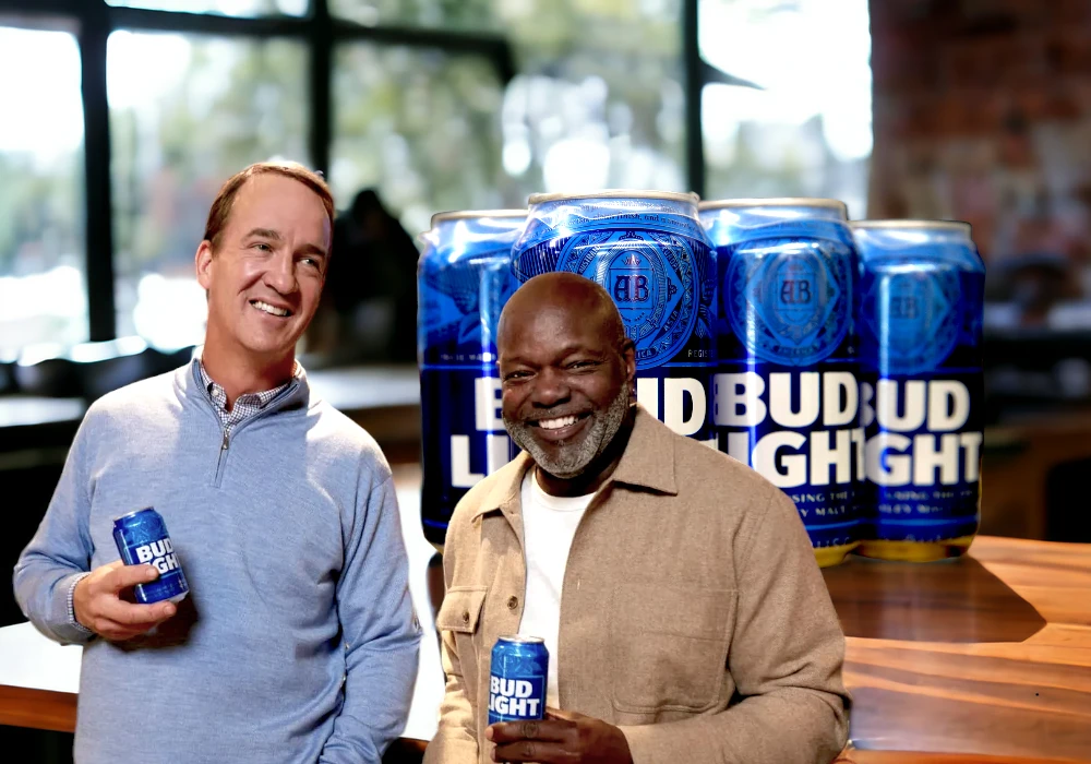 Two men stand smiling while holding Bud Light cans in front of a stack of Bud Light packs. In this clever celebrity endorsement strategy, they are indoors with a large window in the background.