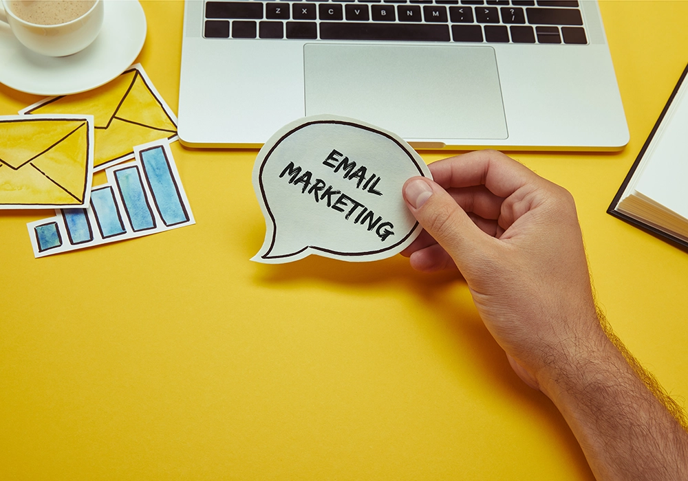 A hand holding a speech bubble with "Email Marketing" written on it next to a laptop, coffee cup, charts, and notebook on a vibrant yellow surface.