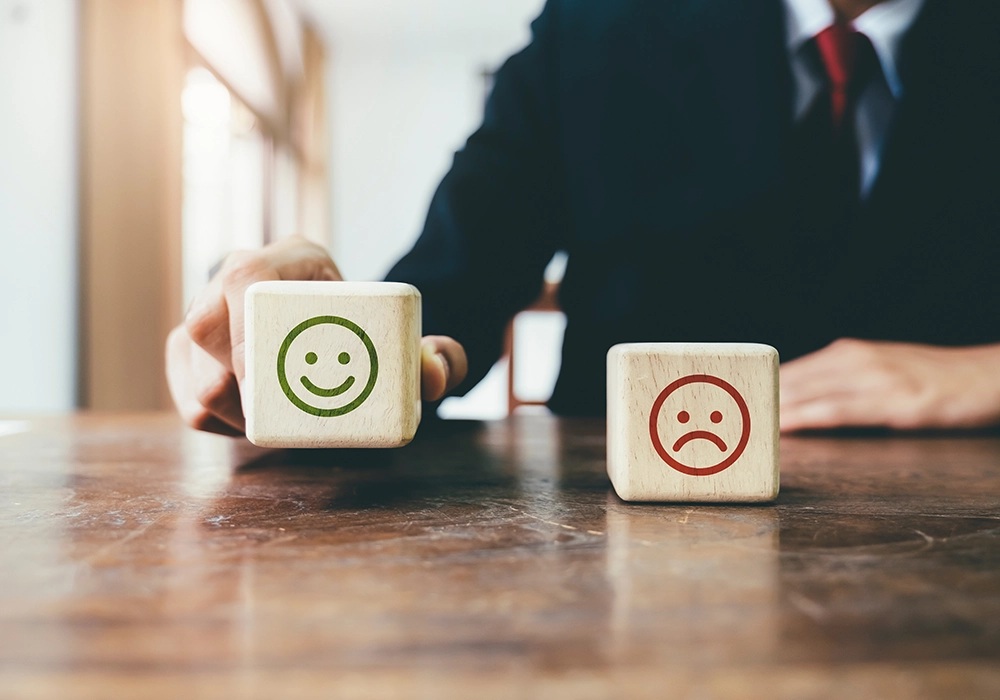 A person in a business suit at a wooden table, flipping a cube with a happy face, while another cube with a sad face is beside it, symbolizing customer review feedback.