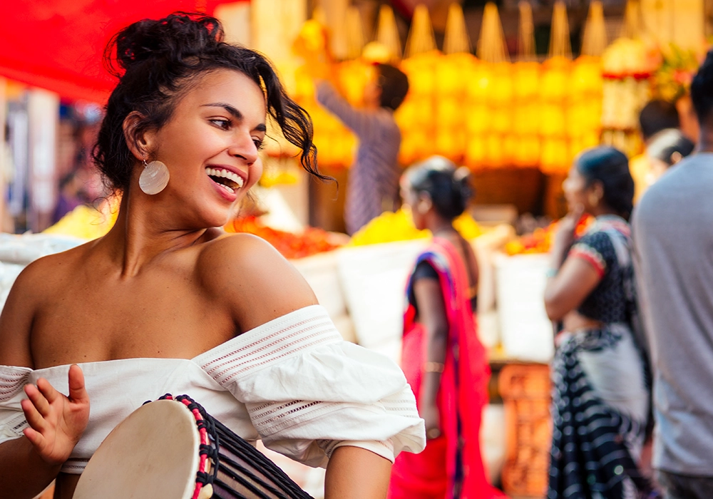 A joyful woman laughing, wearing a white off-the-shoulder top, with a bustling cultural street market scene in the background.