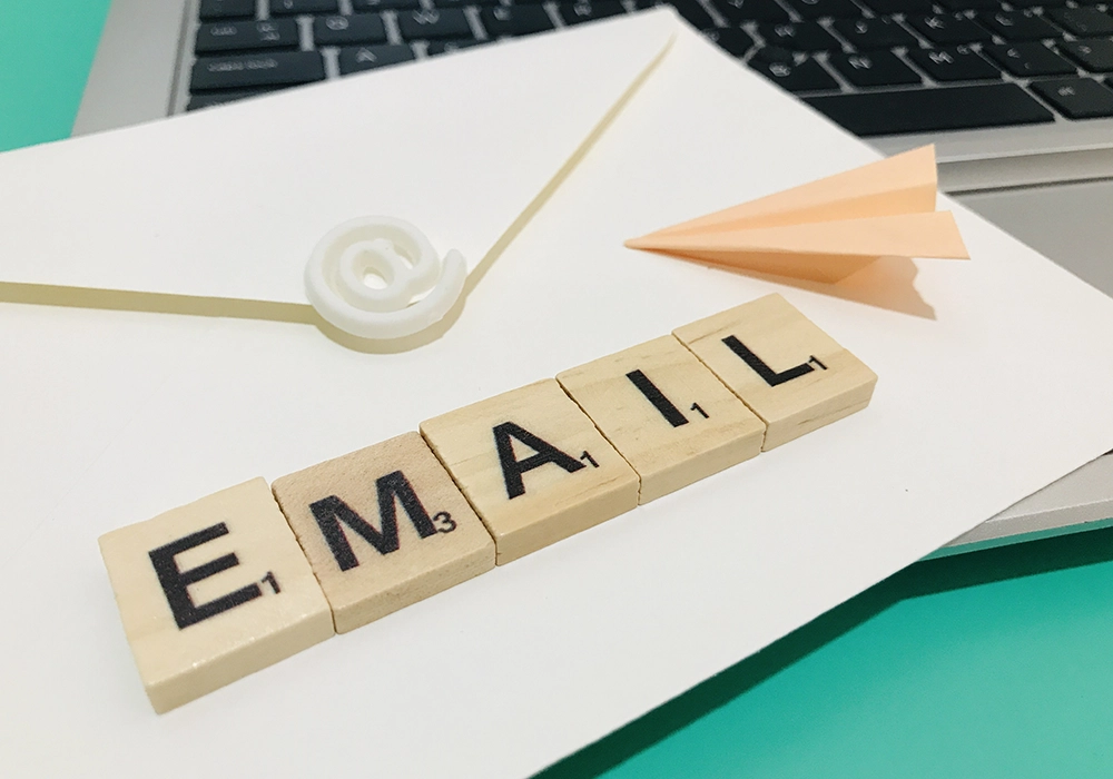 A laptop, an envelope with an email symbol seal, a paper airplane, and Scrabble tiles spelling "EMAIL" are arranged on a light-colored surface, setting the perfect scene for an Email Marketing campaign.