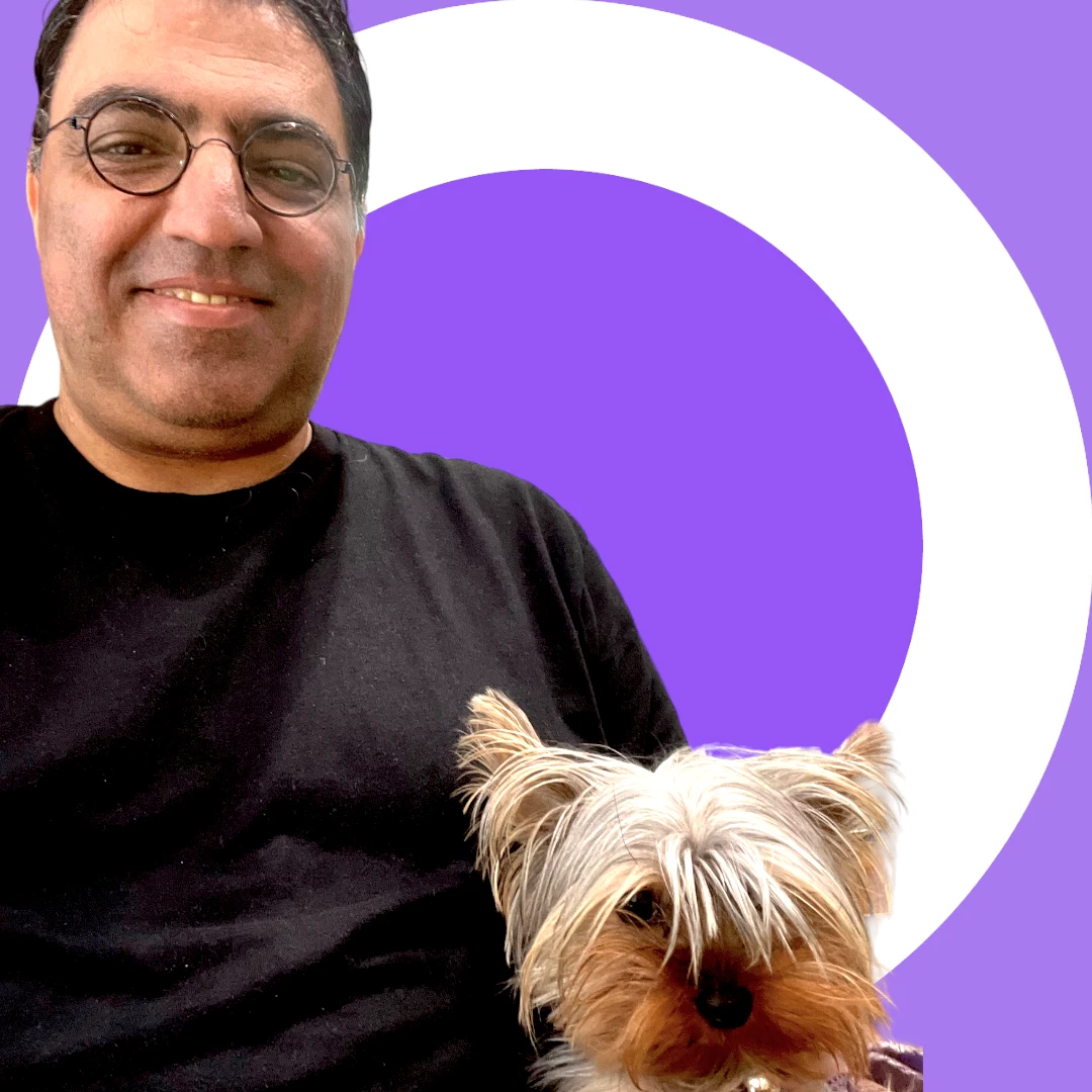 Simon Sarji is wearing glasses and a black shirt smiles while holding a small dog with long fur against a purple background with a white circle.
