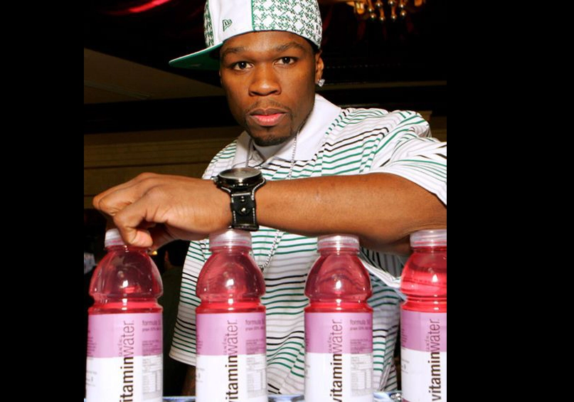 50 Cent in a striped shirt and cap poses with several bottles of Vitamin Water on a table, reminiscent of celebrity food endorsements.