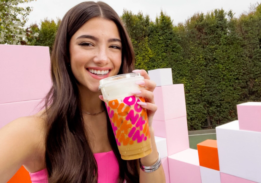 A person with long dark hair smiles while holding a large iced beverage from Dunkin'. They stand outdoors in front of pastel-colored blocks, resembling a scene straight out of one of those glamorous celebrity food endorsements.