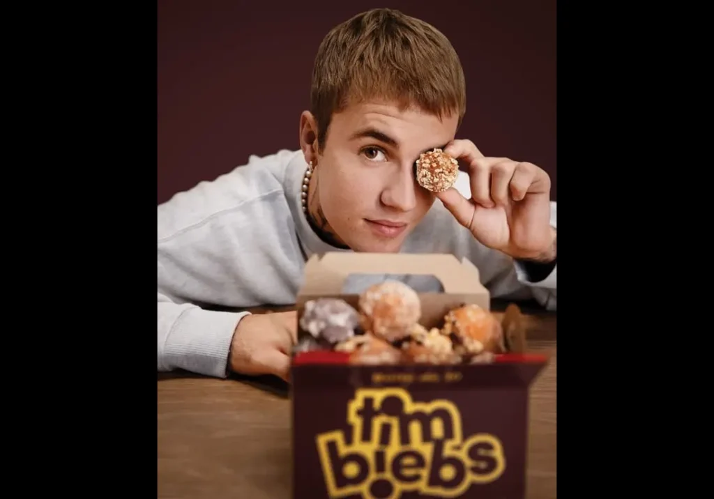 Justin Bieber leans on a table, holding a small round pastry near their eye. On the table is an open box labeled "Timbiebs" filled with various pastries—proof that celebrity food endorsements can be quite delicious.
