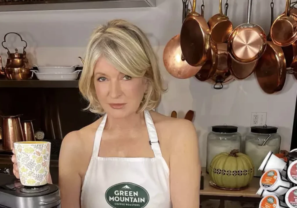 Martha Stewart wearing an apron stands in a kitchen holding a patterned paper cup. Copper pots and kitchen items are in the background, reminiscent of a scene from celebrity food endorsements.