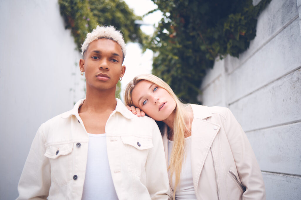 Two individuals stand closely, with one resting their head on the other's shoulder. Both wear light-colored clothing inspired by celebrity fashion collaborations, set against a backdrop of white walls and greenery.