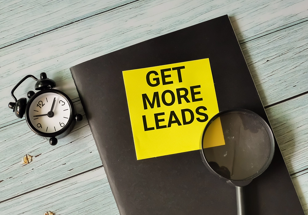 A black notebook with a yellow sticky note that reads "Get More Leads," next to an alarm clock and a magnifying glass on a wooden surface.