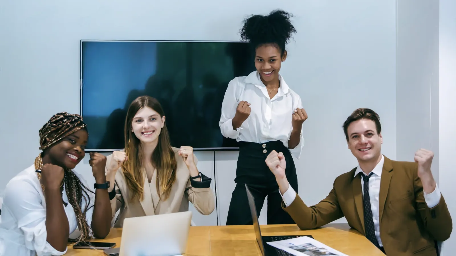 Four people sit and stand around a table with laptops, smiling and raising their fists in a celebratory gesture—a lively scene that could illustrate customer testimonial examples in a modern office meeting room.