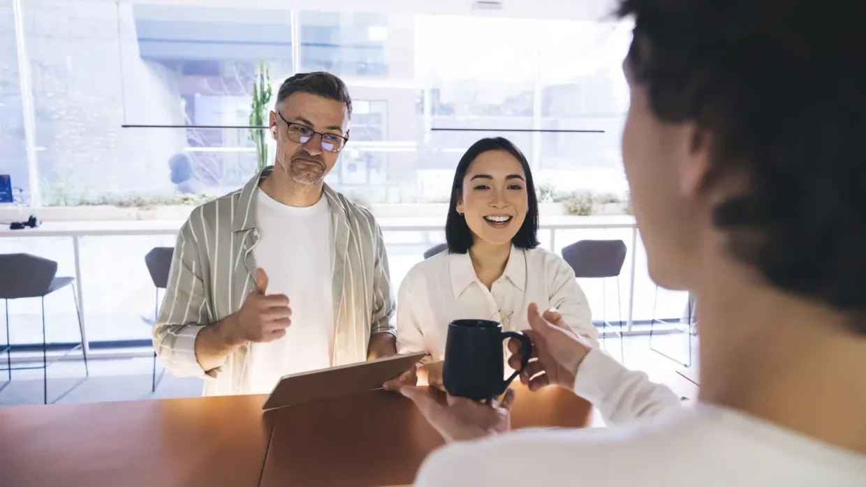 Two people sitting at a table, one giving a thumbs up and the other smiling, while a third person hands over a black mug—capturing a moment of customer experience improvement in action.