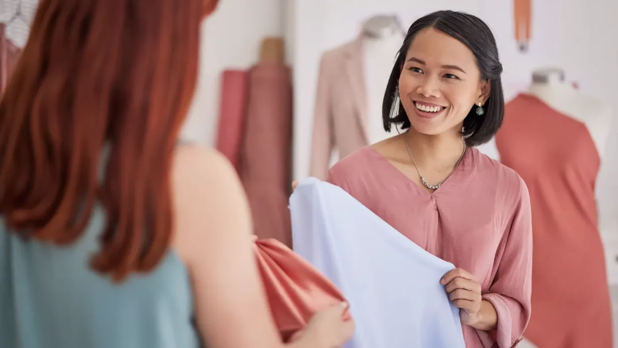 Two women in a clothing store look at fabric swatches together, using customer feedback analysis to guide their choices. One smiles while holding a light blue fabric, and mannequins with dresses are visible in the background.