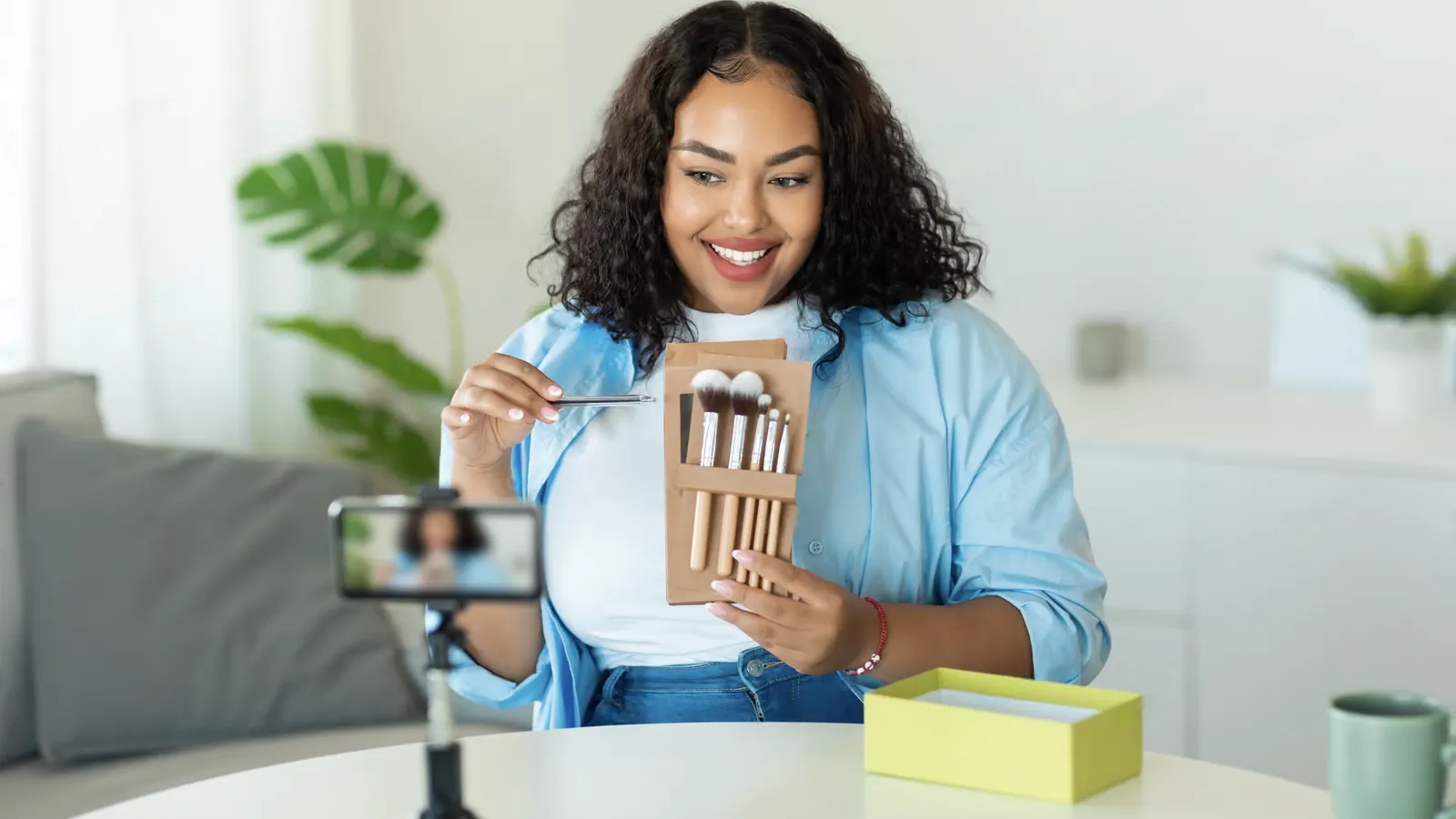 A woman sits on a couch, holding makeup brushes and demonstrating them to a smartphone camera on a tripod—perfectly showcasing influencer marketing strategies in action.