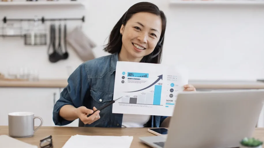 Woman smiling and holding a paper with a rising graph, sitting at a desk with a laptop in a home office setting, reflecting the success of her marketing automation workflow.
