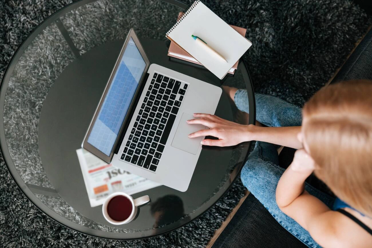 A person uses a laptop at a glass table, exploring Content Writing Tools for SEO, with a notebook, pen, newspaper, and a cup of coffee placed nearby on a dark carpet.