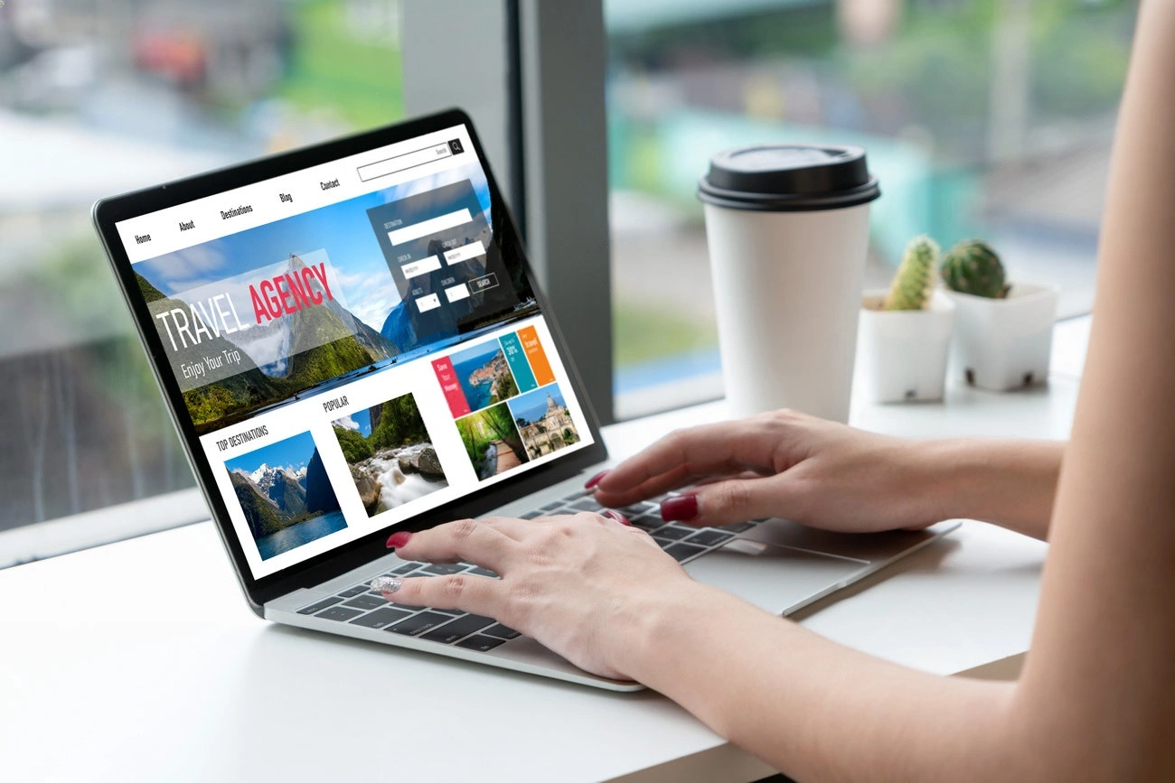 A person using a laptop with a travel agency website open, next to a disposable coffee cup and a small potted cactus by the window—perfect inspiration for anyone who needs a website for their own travel business.