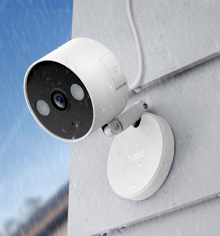 A white outdoor TP-Link Tapo C120 mounted on a wall is wet from rain, with water droplets visible on its surface.
